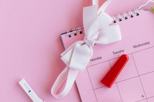 Flat lay of a pregnancy test on a pink background with a calendar and bow.